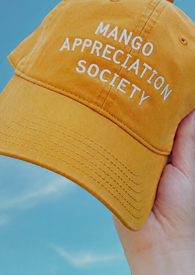 Yellow cap with Mango Appreciation Society text held up against a blue sky.